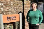 Lisa stands next to a brick wall with an orange sign which says Amber Farm Place in navy text. 